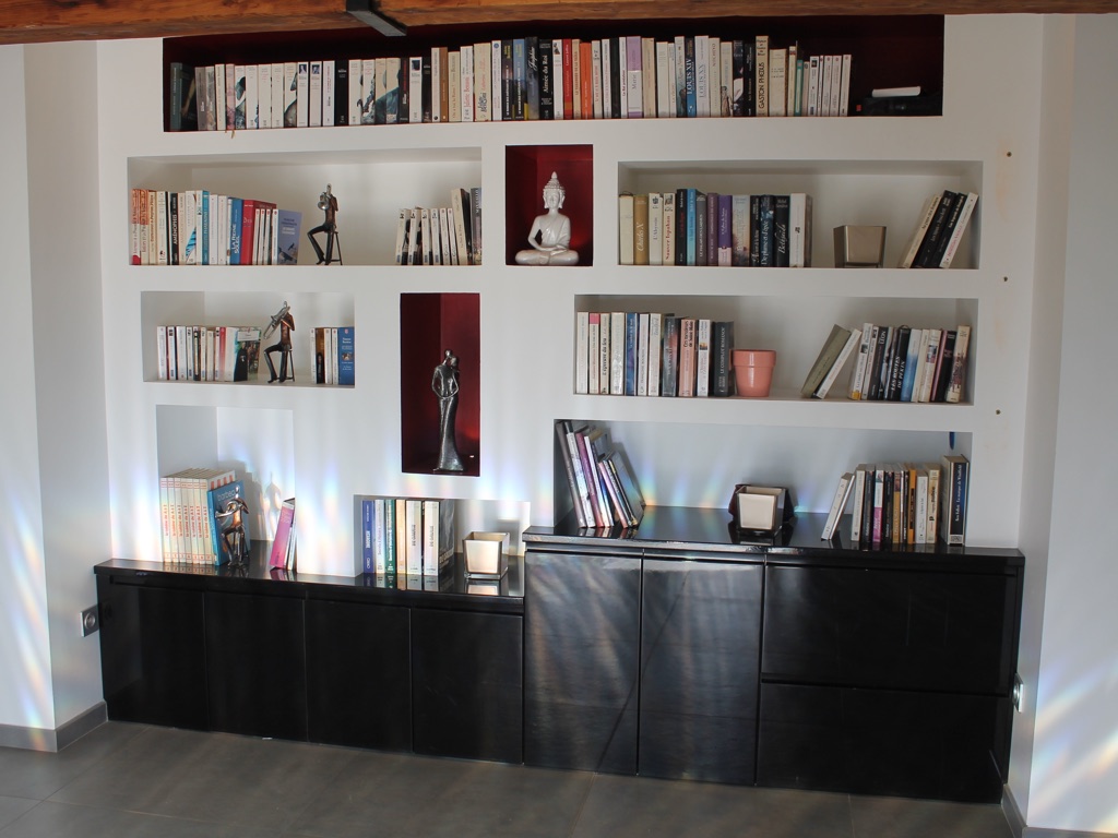 Bibliothèque moderne encastrée avec niches murales blanches et accents rouges. Meuble de rangement bas laqué noir brillant décoré de livres et d'un Bouddha.