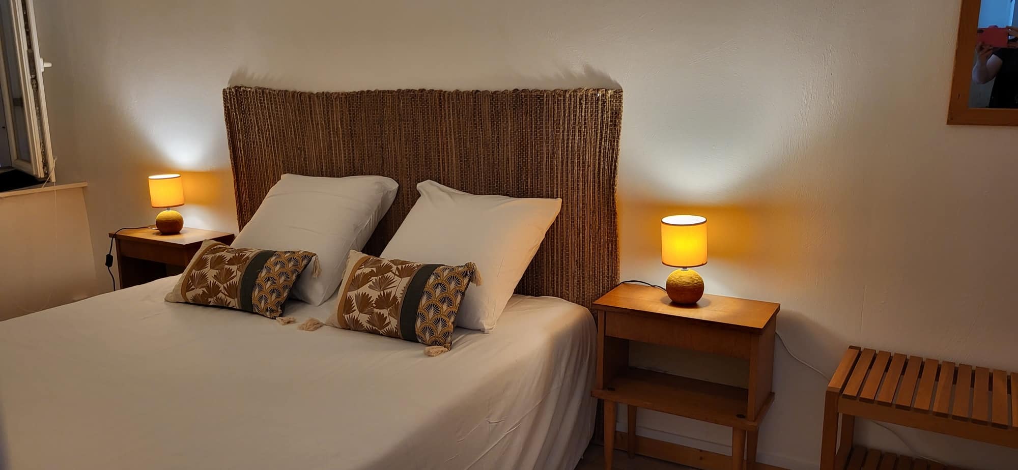Chambre cozy avec lit double, tête de lit en rotin naturel, et deux veilleuses à abat-jour orange créant une ambiance chaleureuse.