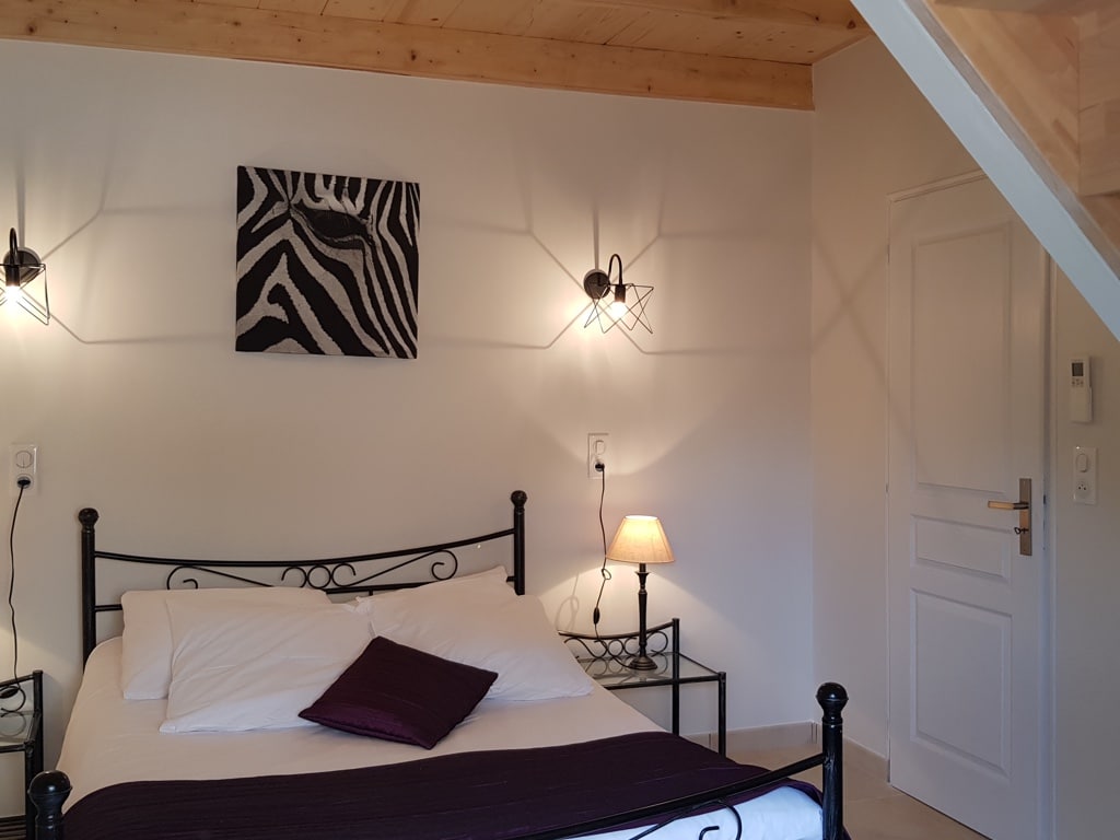 Lit double à tête en fer forgé, literie blanche et jeté prune. Toile murale zèbre audacieuse entre deux appliques design noires. Plafond bois.