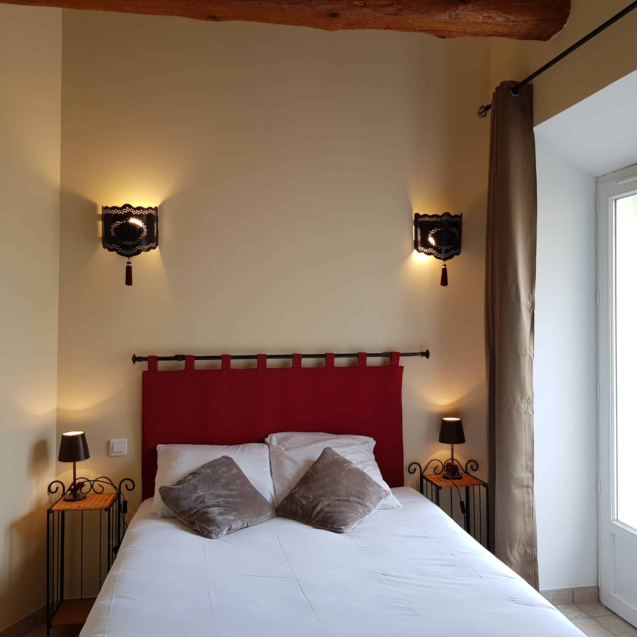 Lit double avec tête de lit matelassée rouge. Éclairage chaud par deux appliques marocaines noires et tables de chevet en fer forgé.