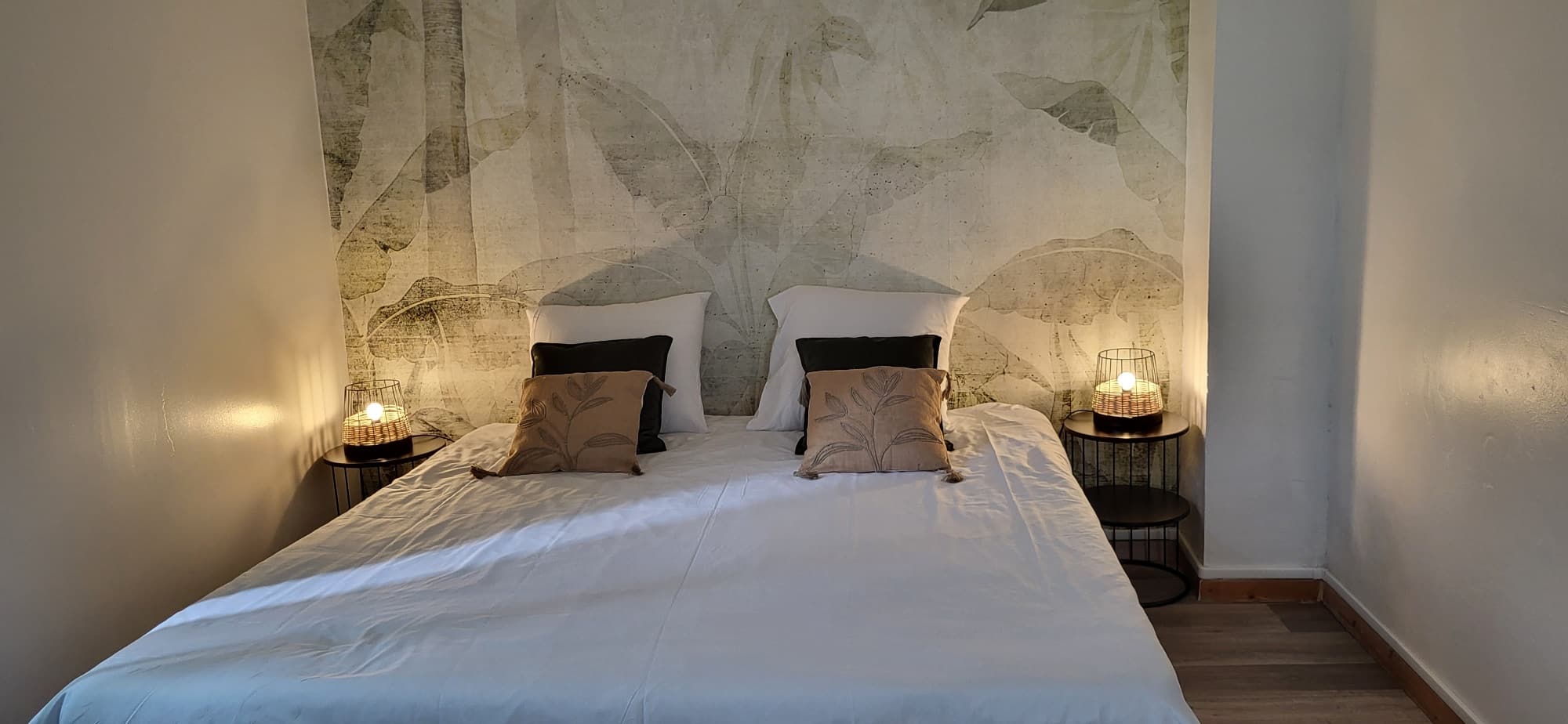 Chambre moderne : lit double blanc devant un grand mural tropical pâle. Deux tables de chevet noires avec lampes tressées à lumière chaude.