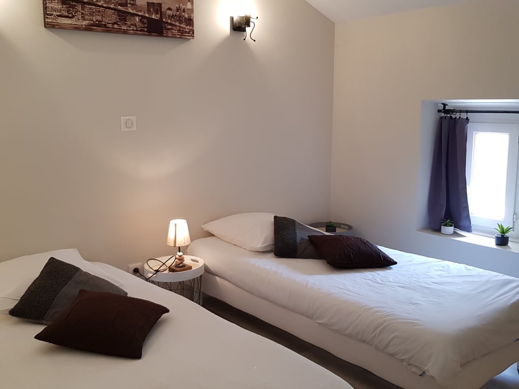 Chambre moderne lumineuse : deux lits jumeaux blancs, oreillers sombres, table de chevet, lampe allumée et tableau New York. Fenêtre cosy.