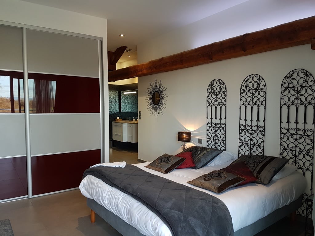 Chambre cozy: lit confortable, grande tête de lit en fer forgé. Poutre en bois rustique. Dressing coulissant rouge et gris vers la SDB.