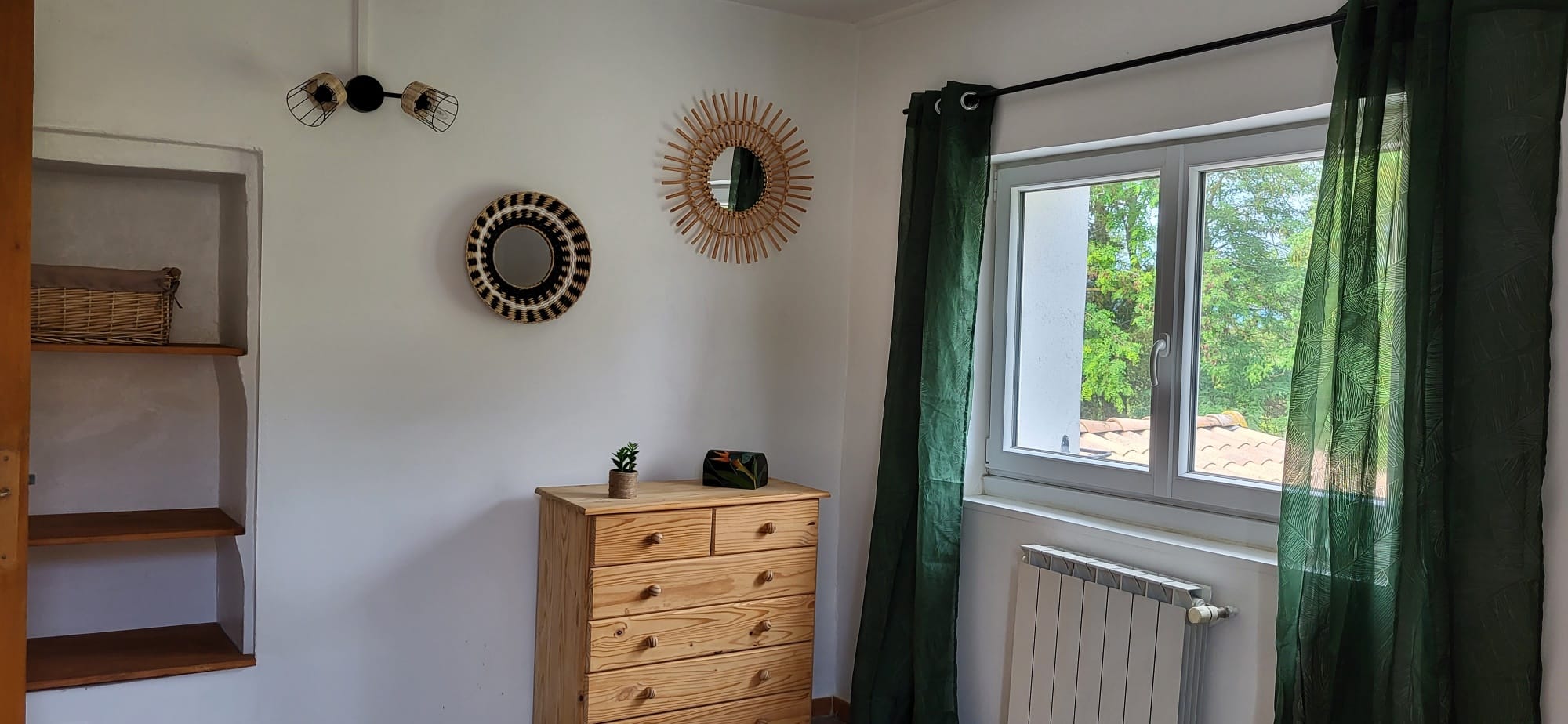 Commode en pin clair et deux miroirs ronds en rotin sur mur blanc. Décoration bohème avec rideaux verts à motifs et niche.