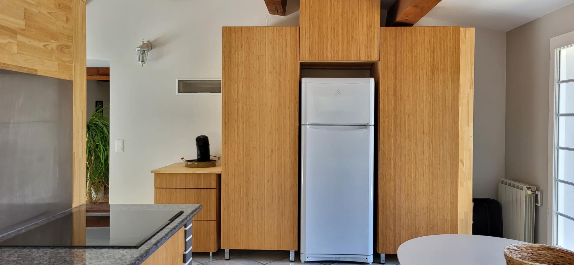 Cuisine moderne avec armoires en bois clair de bambou encastrées autour d'un réfrigérateur Indesit. Comptoir en granit noir à gauche.