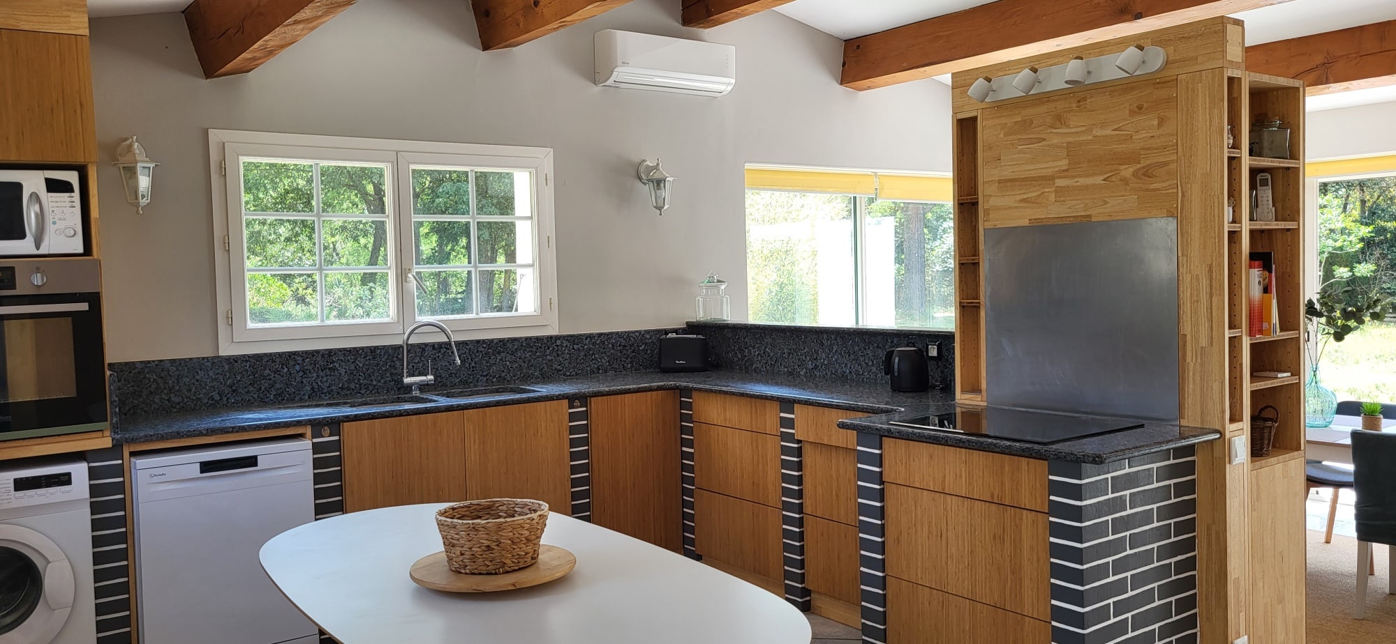 Lumineuse cuisine moderne/rustique. Meubles en bois clair, granit foncé, îlot en brique noire et étagères. Grandes fenêtres sur la nature.