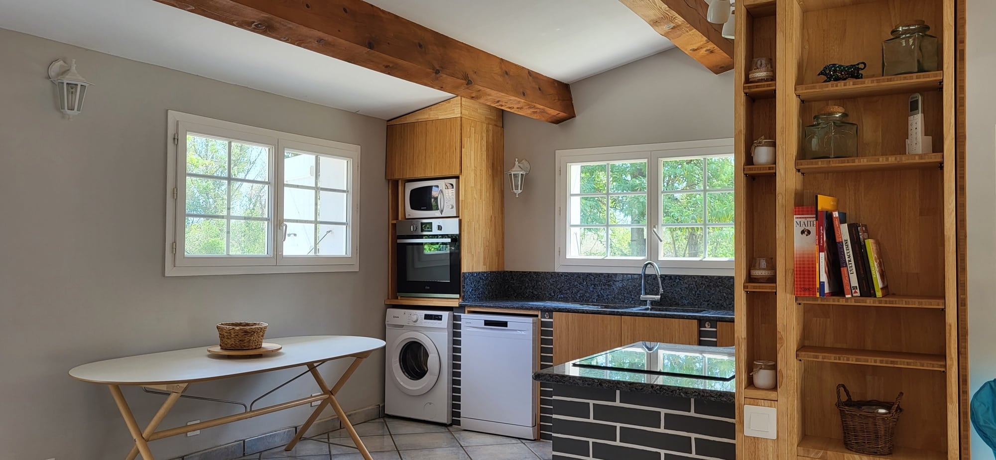 Cuisine rustique moderne lumineuse. Armoires en bois clair, poutres apparentes, îlot noir en briques et plan de travail en granit.