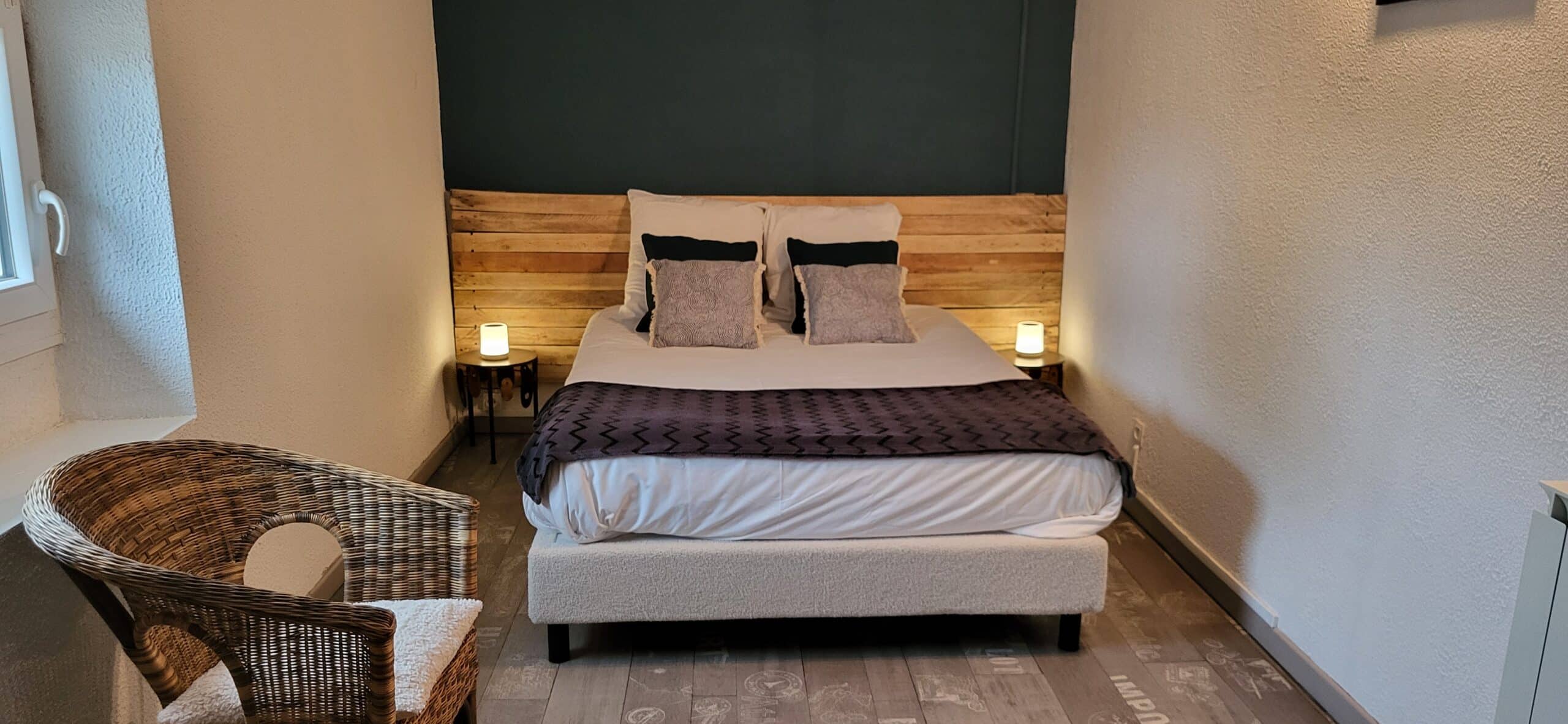 Chambre cosy avec lit double, tête de lit en bois, mur bleu canard et fauteuil en rotin aux lampes tamisées.