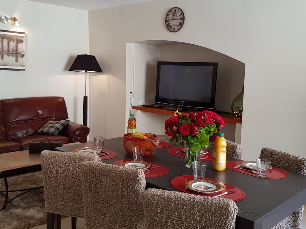 Salon et salle à manger avec canapé en cuir rouge, table en bois sombre, chaises texturées, bouquet de roses et télévision encastrée sous une horloge murale.