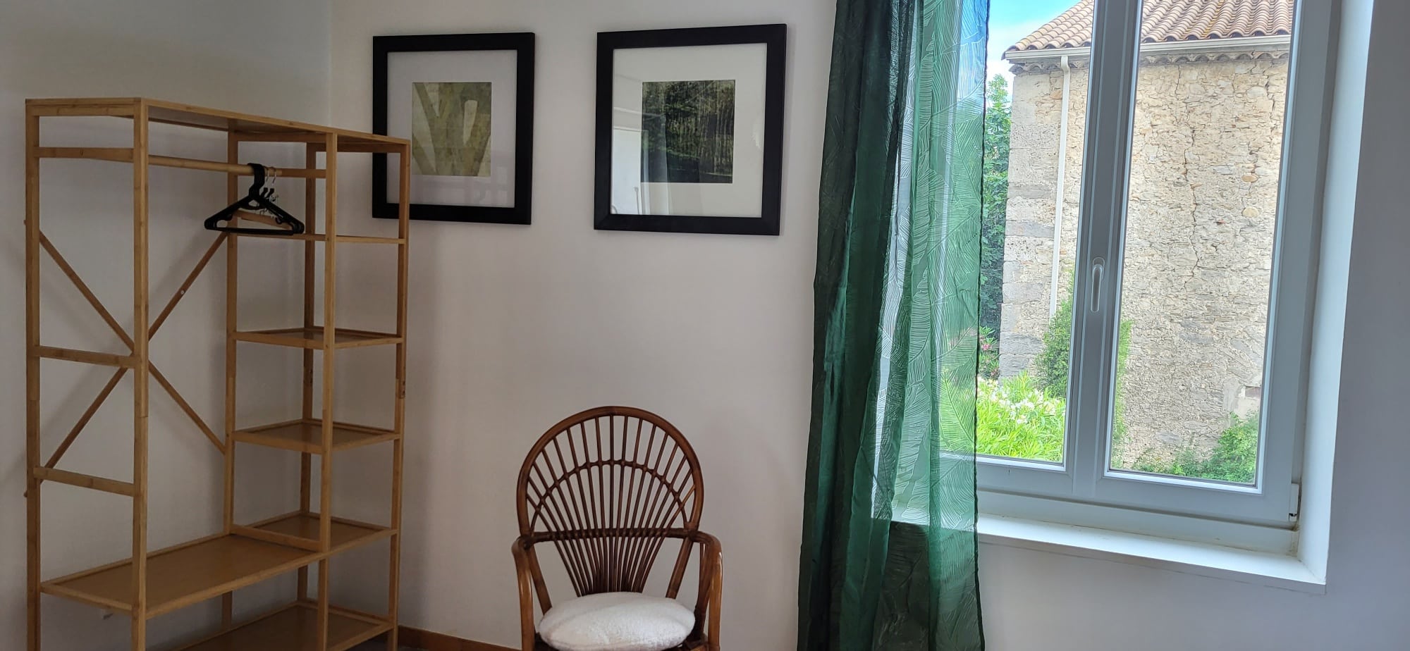 Intérieur moderne avec penderie en bambou, fauteuil en rotin, décorations murales et fenêtre avec rideau vert devant un bâtiment en pierre.