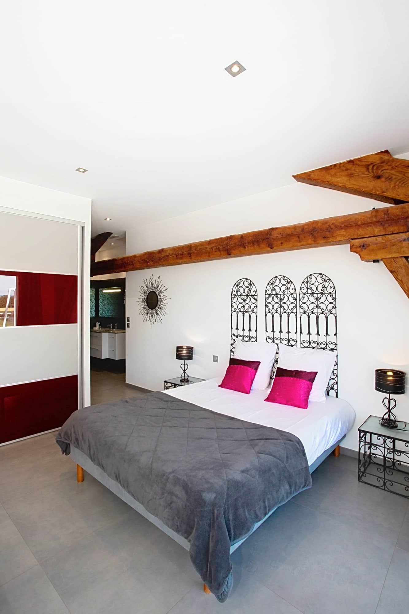Chambre moderne avec poutres apparentes, lit gris, coussins fuchsia et tête de lit en fer forgé noir.
