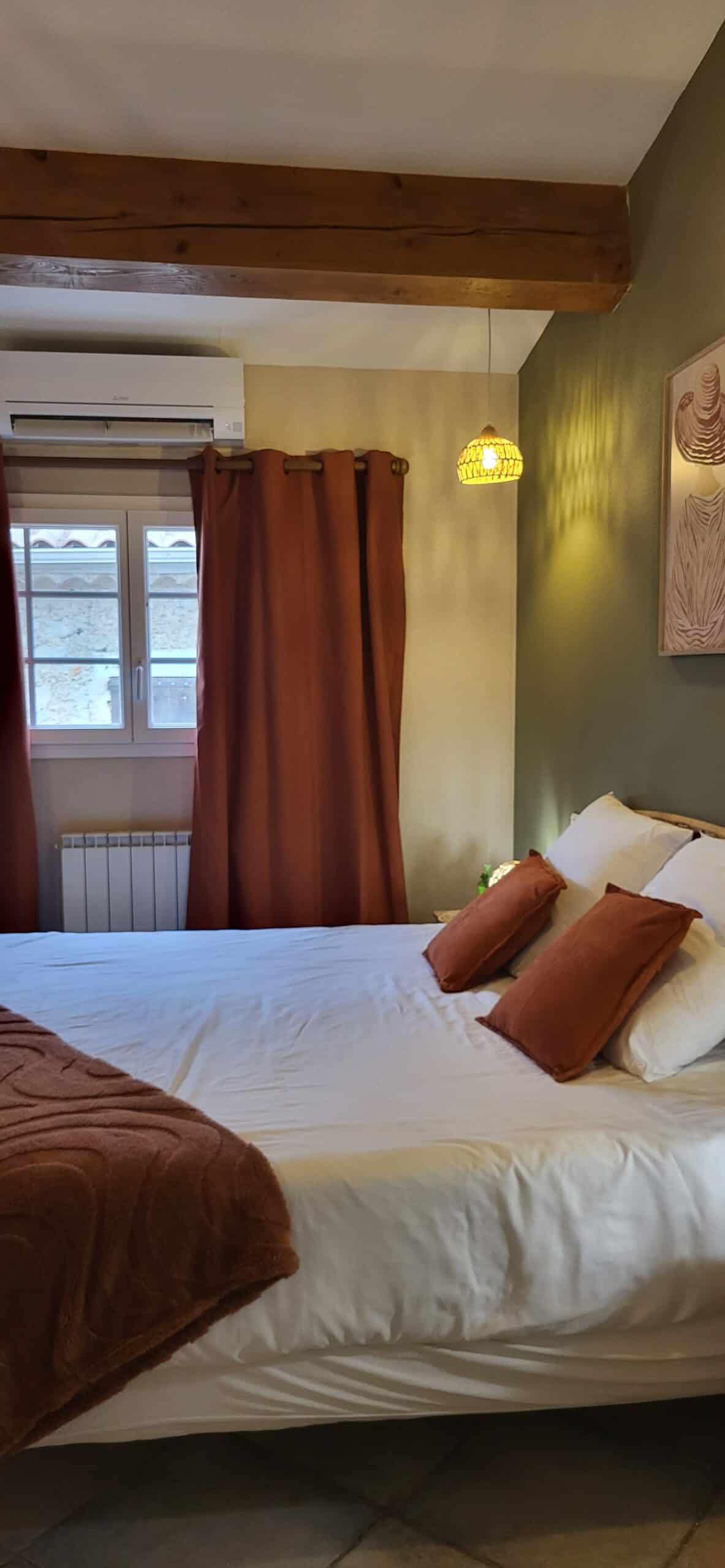 Chambre avec lit blanc, coussins rouille, poutre apparente, rideaux terracotta et mur vert olive chaleureux.