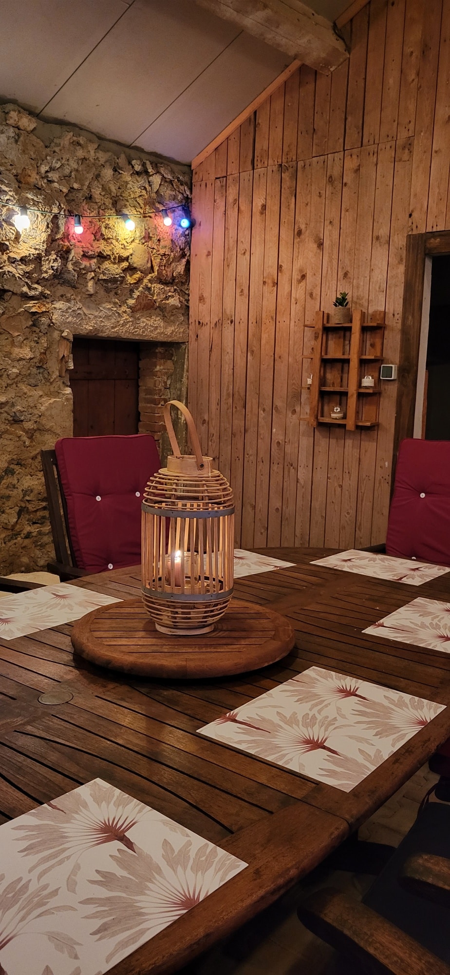 Ambiance dîner rustique sur terrasse. Lanterne en bambou au centre d'une table en bois. Murs en pierre ancienne et bardage bois, guirlande lumineuse colorée.