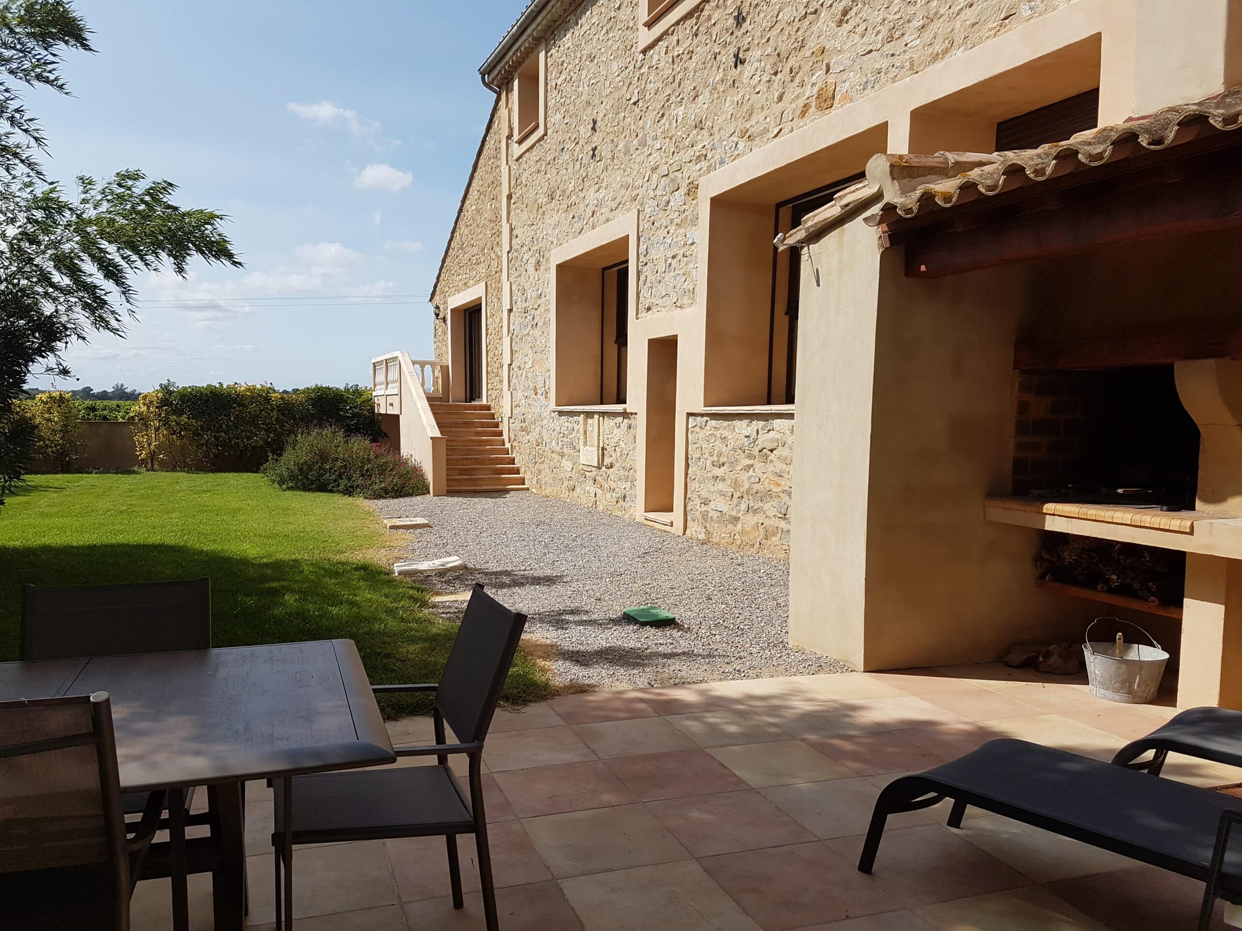 Patio aménagé devant une maison traditionnelle en pierre ocre. Il y a un barbecue extérieur, une table de jardin et des escaliers menant à l'étage.