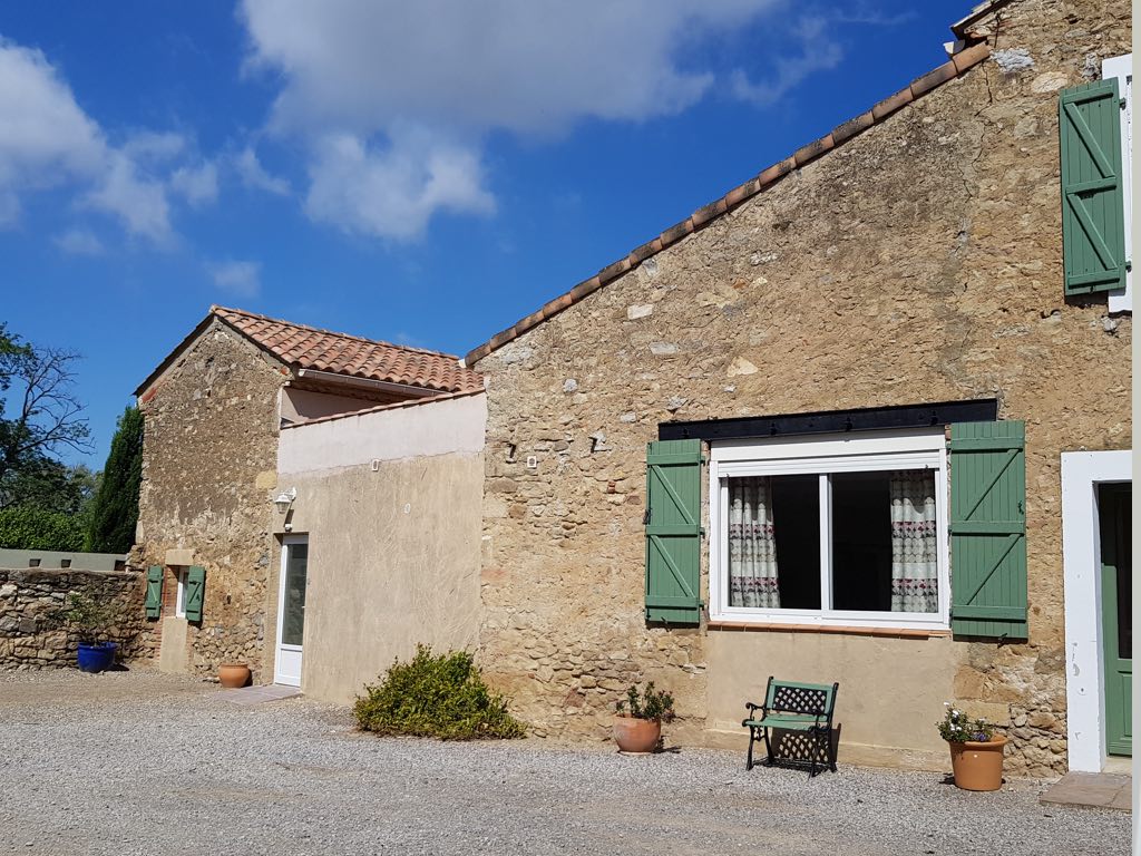 Mas traditionnel provençal avec murs en pierre sèche et toits en tuiles rouges. Façade ensoleillée aux volets verts et chaise décorative sur gravier.