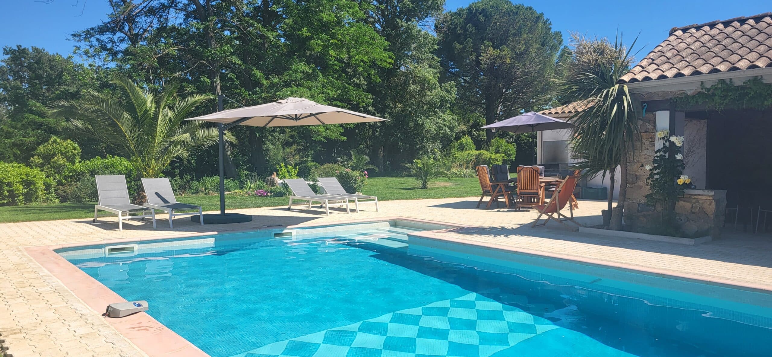 Piscine bleue avec transats, parasols et terrasse ensoleillée dans un jardin arboré avec salon de jardin et pool house.