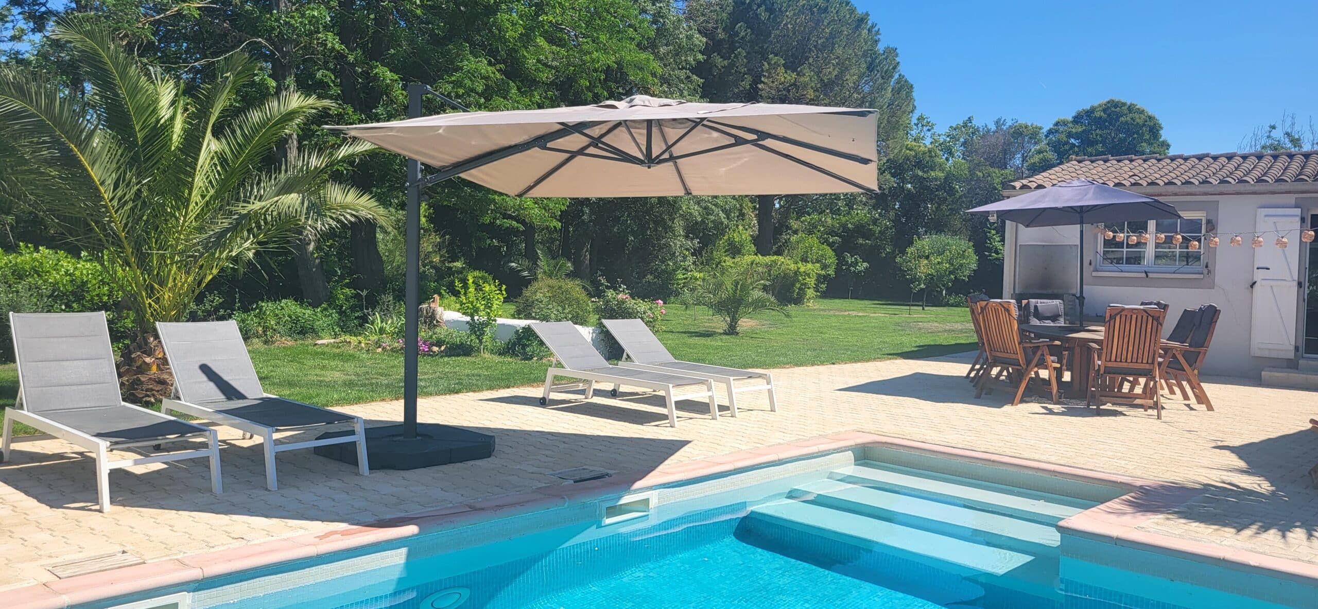 Piscine avec terrasse, chaises longues sous un grand parasol déporté et coin repas dans un jardin verdoyant.