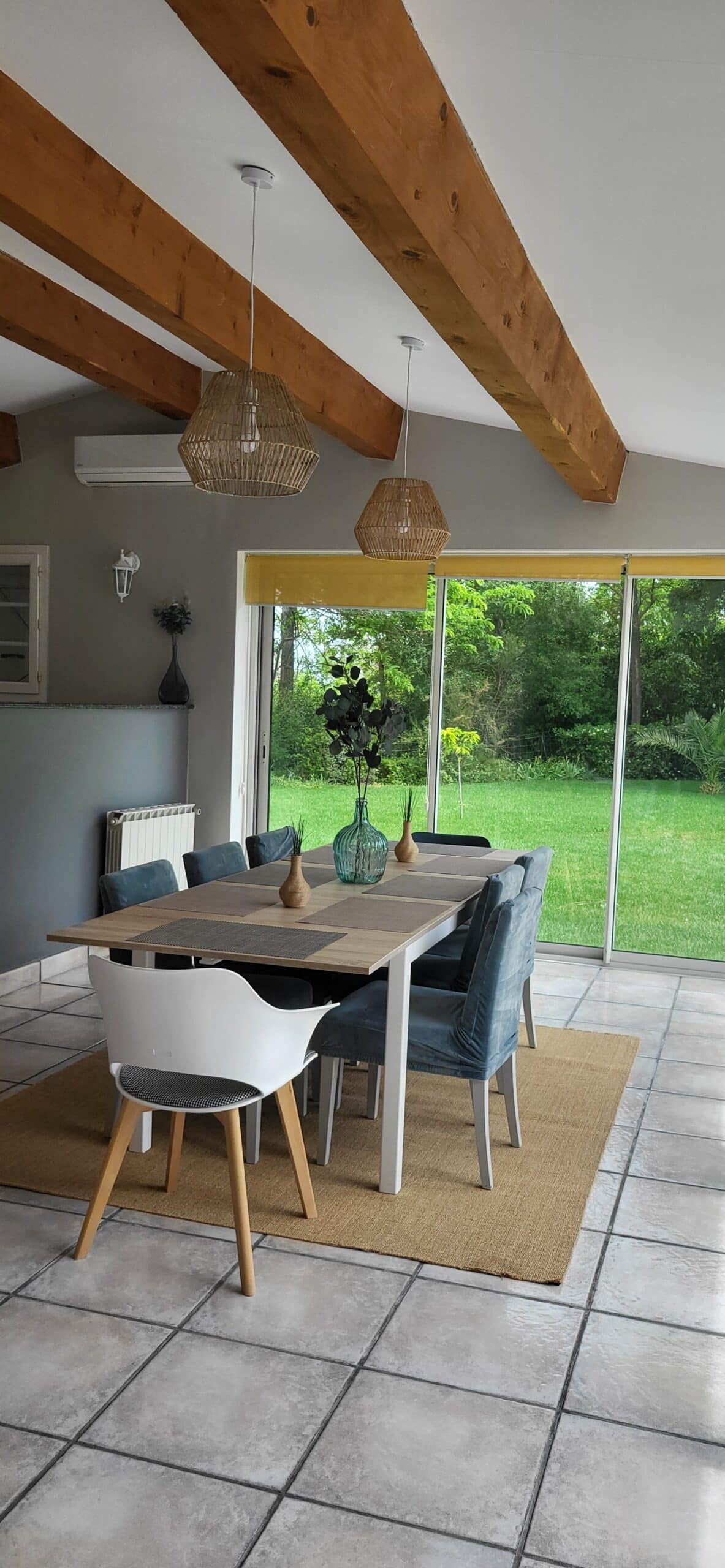 Salle à manger avec poutres apparentes, table en bois, chaises en velours bleu et suspensions en osier sur jardin.