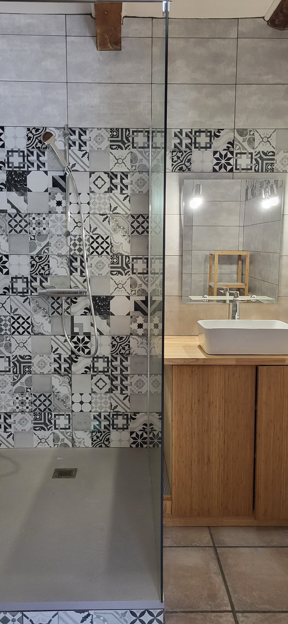 Douche italienne avec carrelage à motifs géométriques gris. Vasque blanche sur meuble en bambou et miroir éclairé.