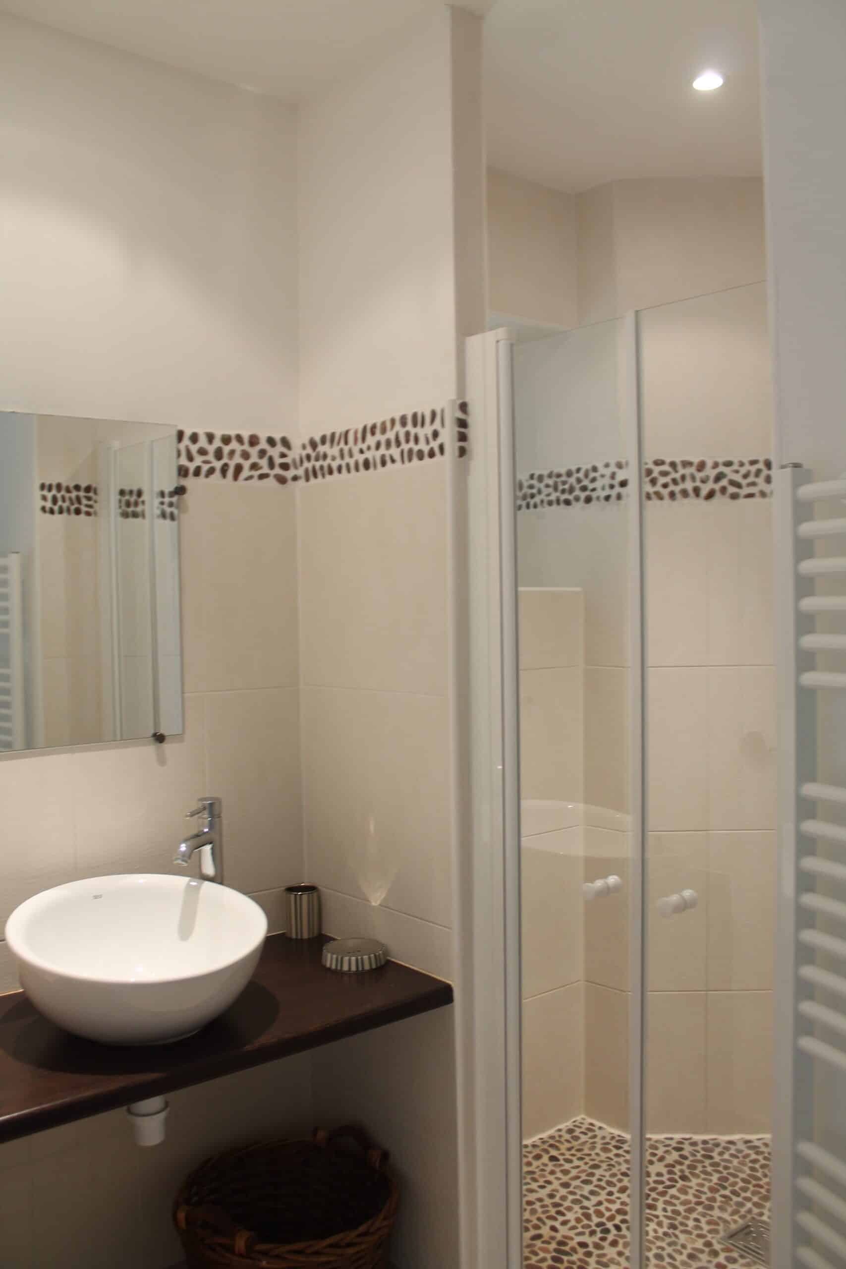 Salle de bain moderne. Vasque blanche sur plan bois foncé, carrelage beige et miroir. Douche avec paroi vitrée et sol en galets naturels.
