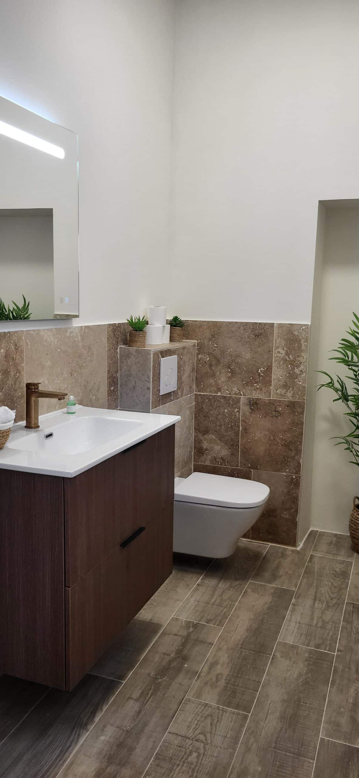 Salle de bain moderne avec meuble en bois rainuré, vasque blanche, robinet doré et WC suspendu blanc.