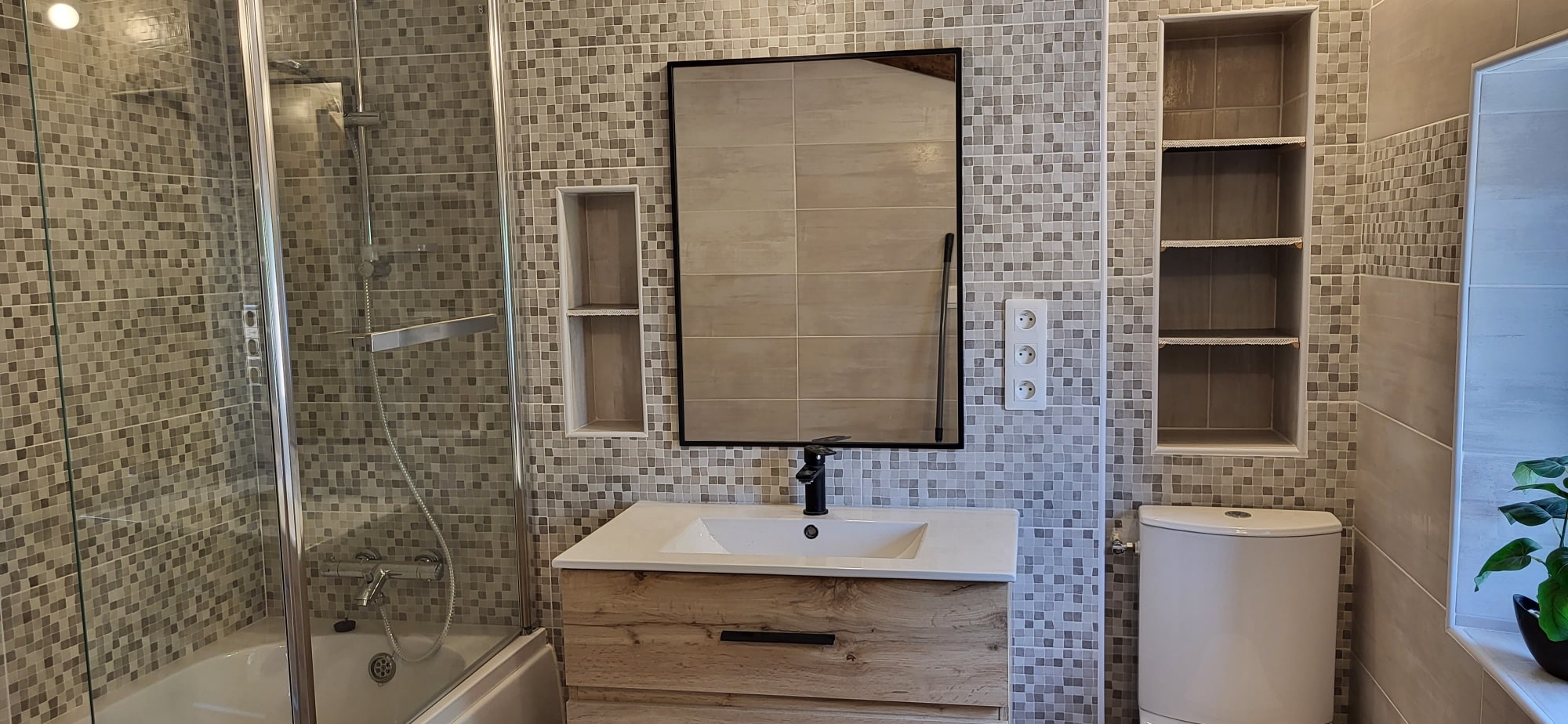 Rénovation de salle de bain moderne. Meuble vasque bois, miroir noir, douche/baignoire. Mur en mosaïque gris-beige avec niches encastrées.