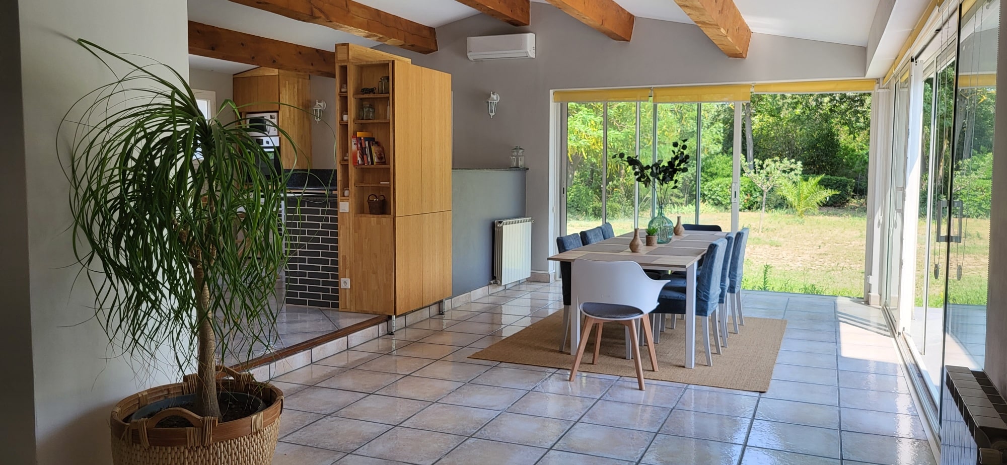 Salle à manger moderne et lumineuse avec table pour 8 sur tapis. Cuisine ouverte en bois, plante verte luxuriante et véranda avec vue jardin.