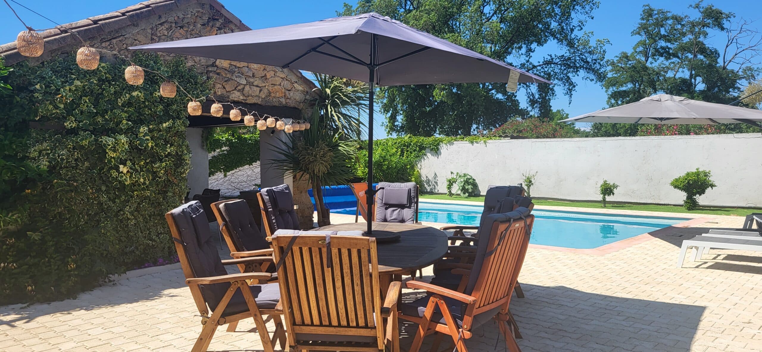 Salon de jardin en bois sous parasol gris, terrasse avec piscine et mur de pierre aux guirlandes.