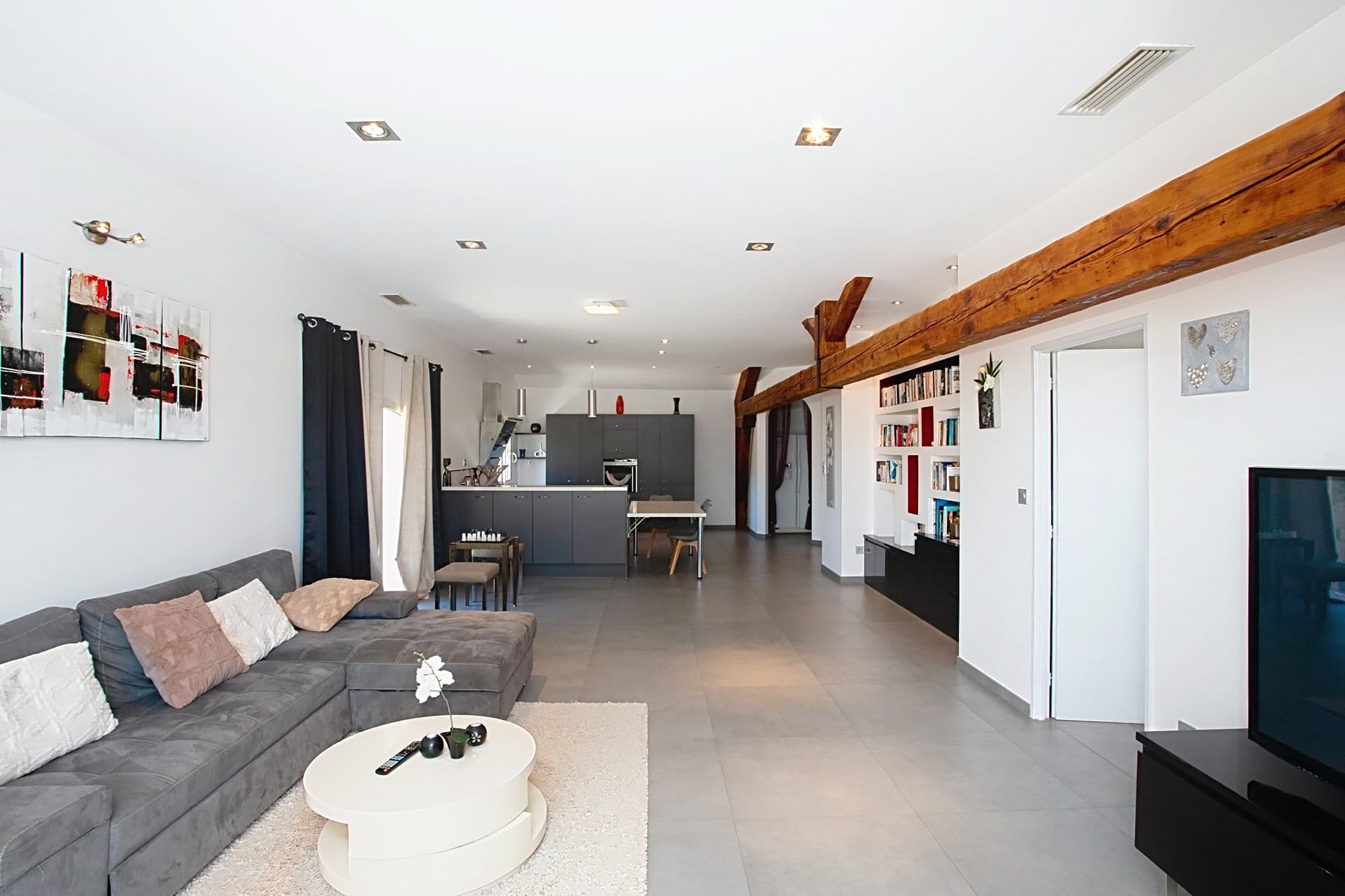Salon moderne avec canapé gris, poutre en bois apparente et cuisine ouverte sur un carrelage gris clair.
