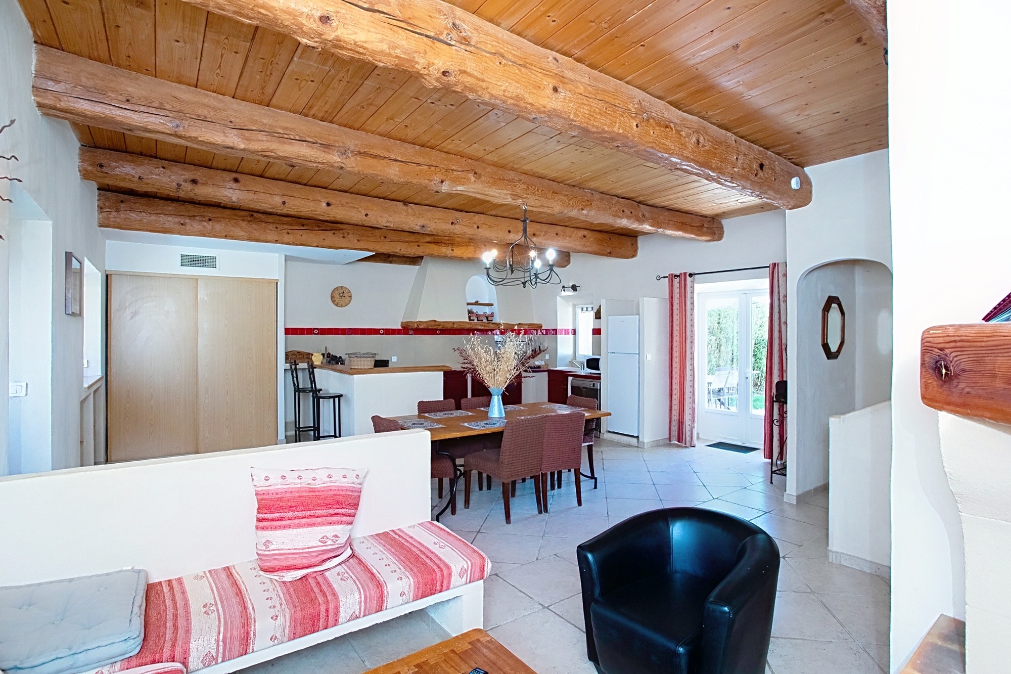 Salon rustique aux poutres apparentes avec salle à manger, cuisine ouverte et canapé aux motifs rouges.