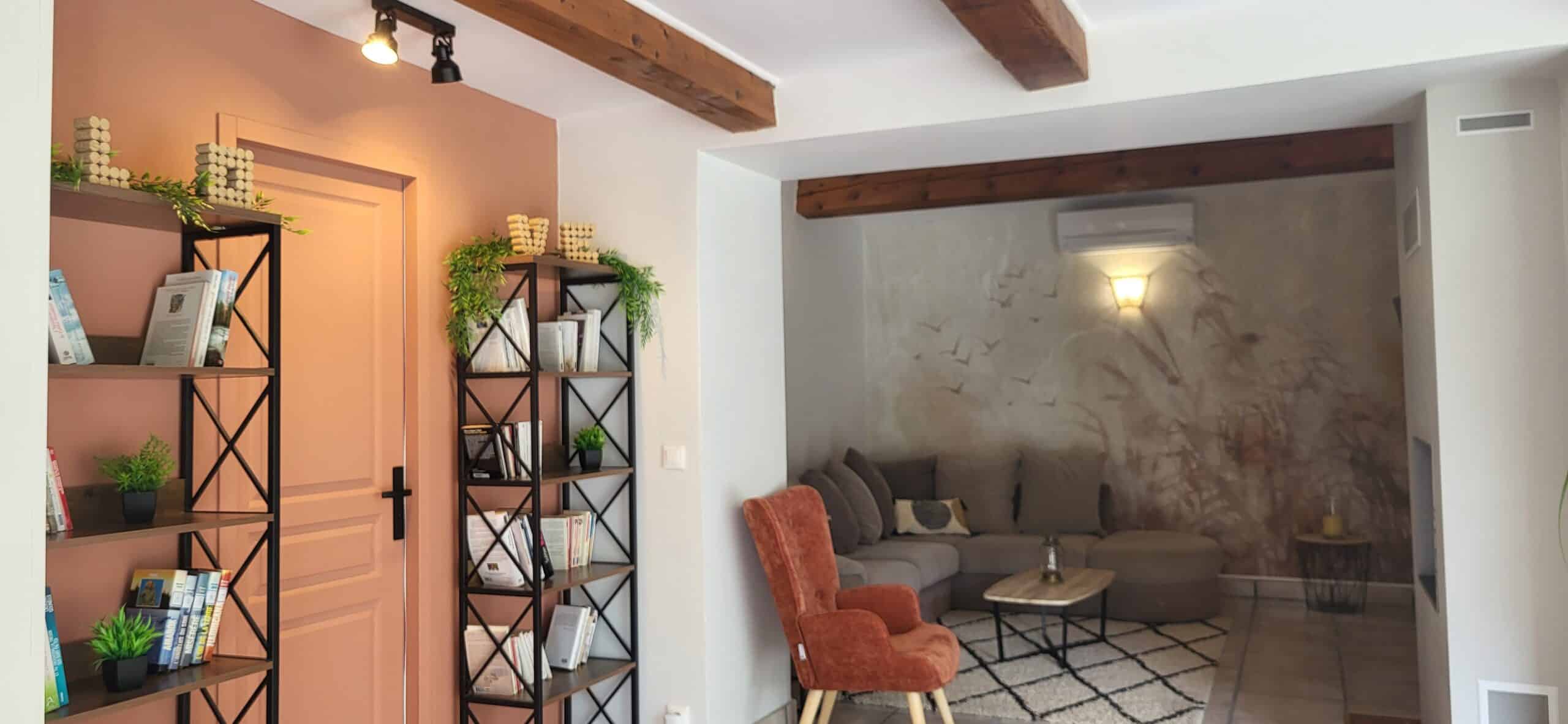 Salon cosy avec mur terracotta, étagères noires, fauteuil en velours orange et canapé gris sous des poutres en bois.