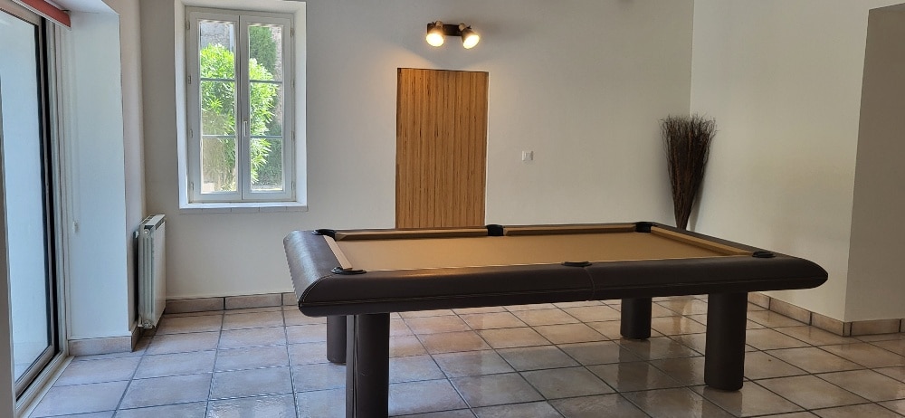 Intérieur moderne : table de billard marron, sol carrelé, fenêtre lumineuse et porte en bois texturé sous une applique murale.