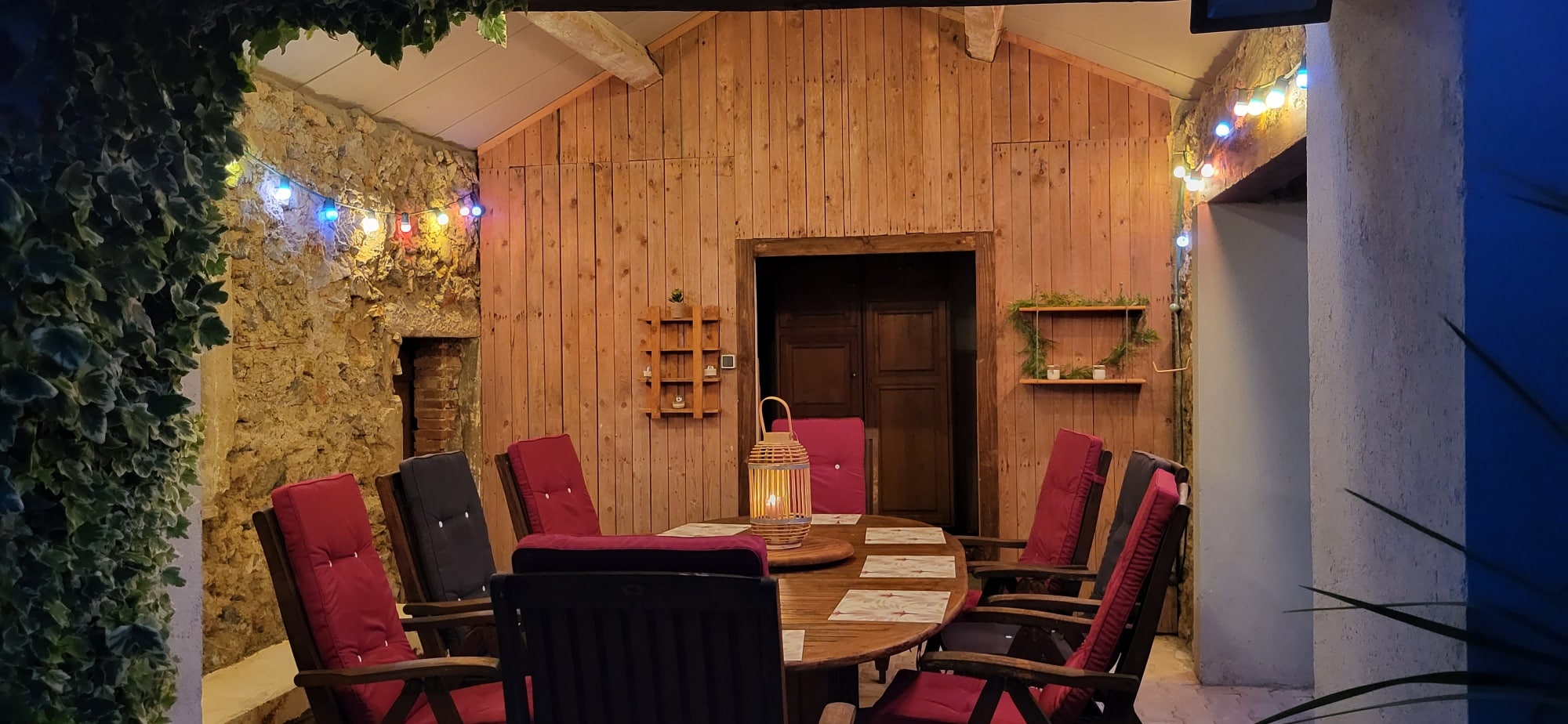 Terrasse chaleureuse pour dîner: grande table en bois, chaises rouges, mur en pierre et lierre. Ambiance tamisée par guirlandes et lanterne.