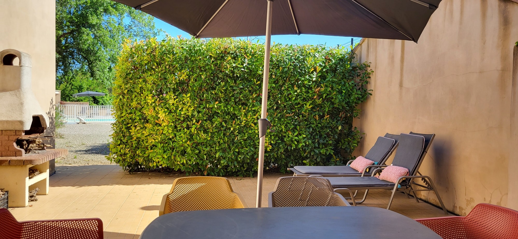 Terrasse ensoleillée avec parasol, chaises longues, table de jardin et haie de verdure. Barbecue en pierre visible.
