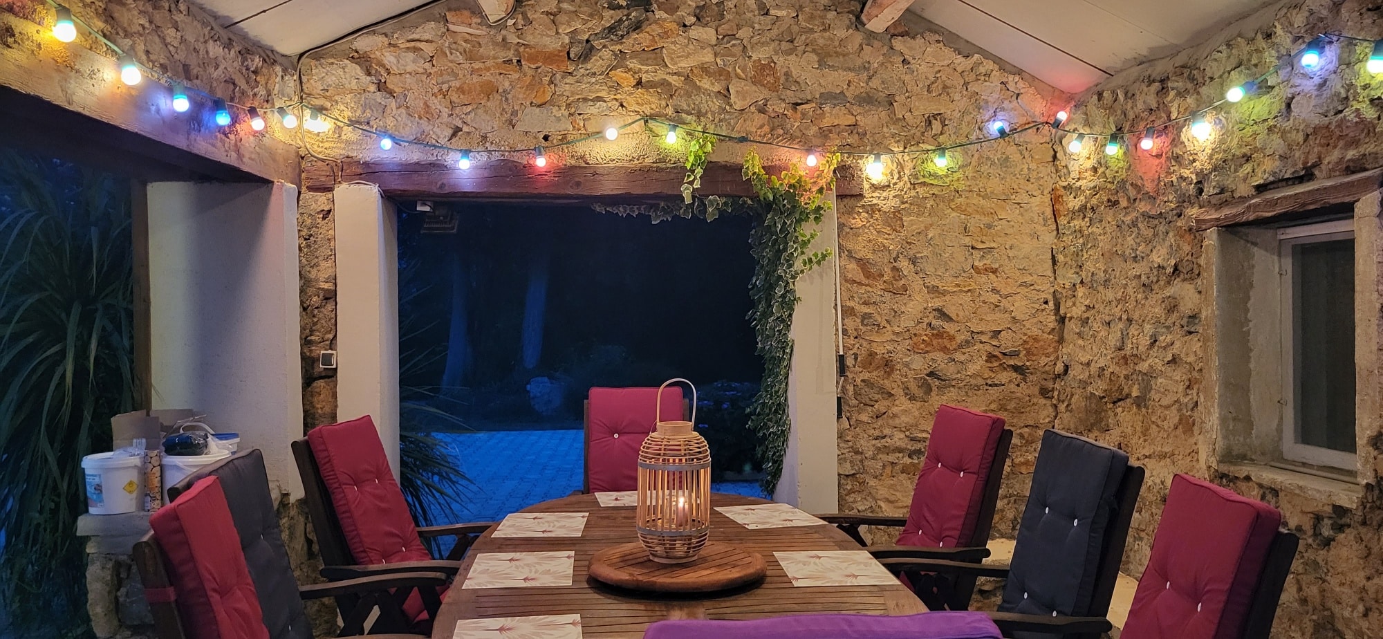Terrasse couverte en pierre brute. Guirlande lumineuse colorée au-dessus d'une table en bois pour le dîner. Coussins rouges et lanterne en osier.