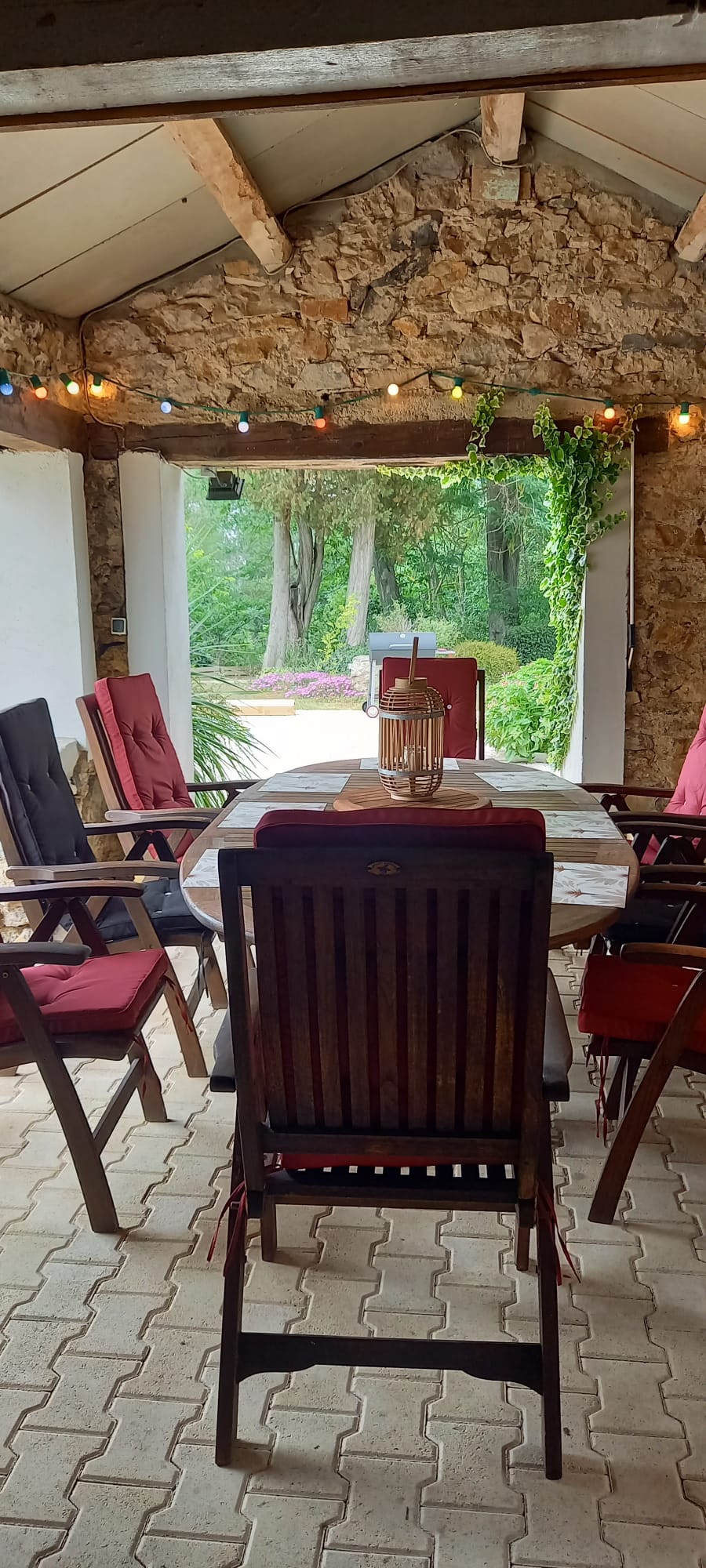 Terrasse couverte rustique avec murs en pierre, guirlande lumineuse. Table en bois et chaises rouges donnant sur un jardin verdoyant.