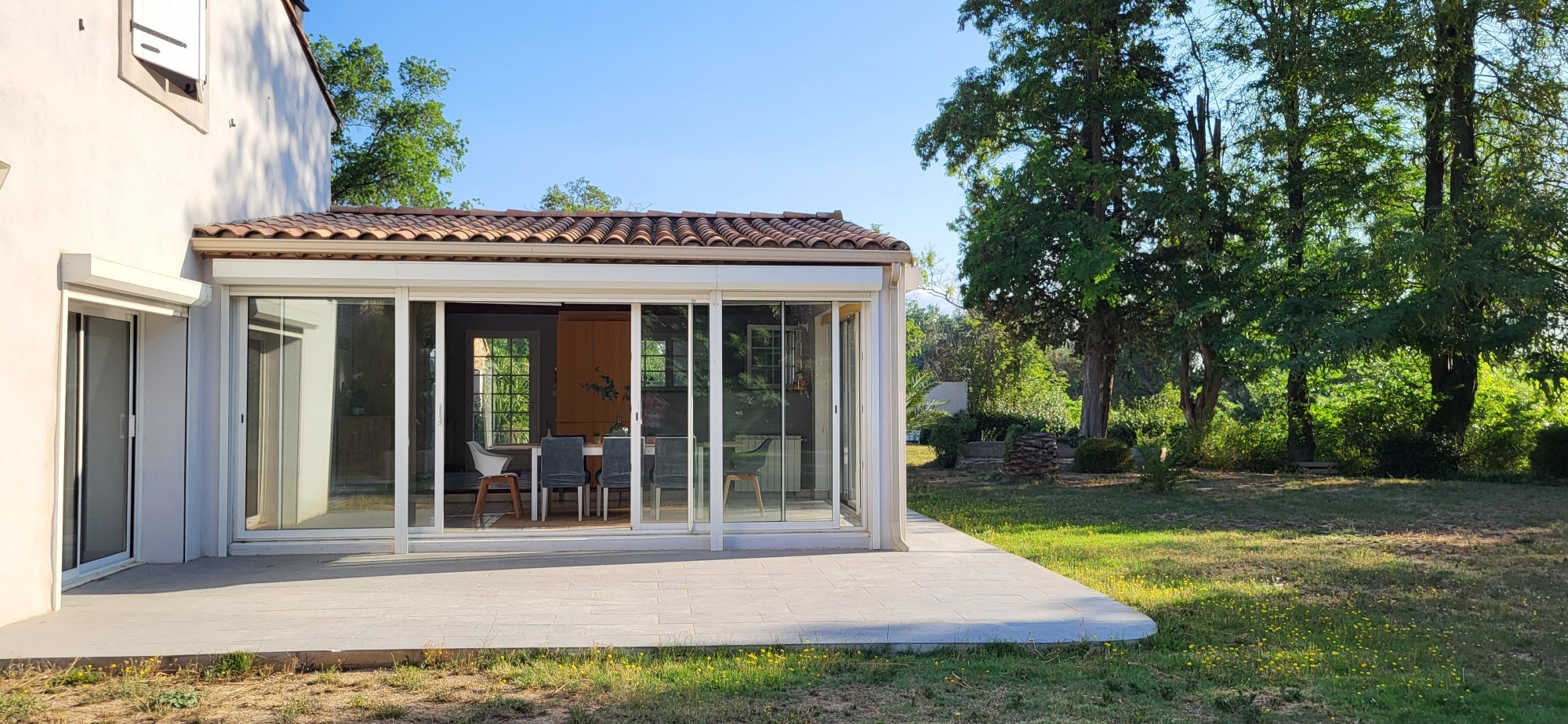 Lumineuse véranda moderne blanche avec baies vitrées et toit en tuiles. Elle s'ouvre sur une grande terrasse et un jardin arboré ensoleillé.