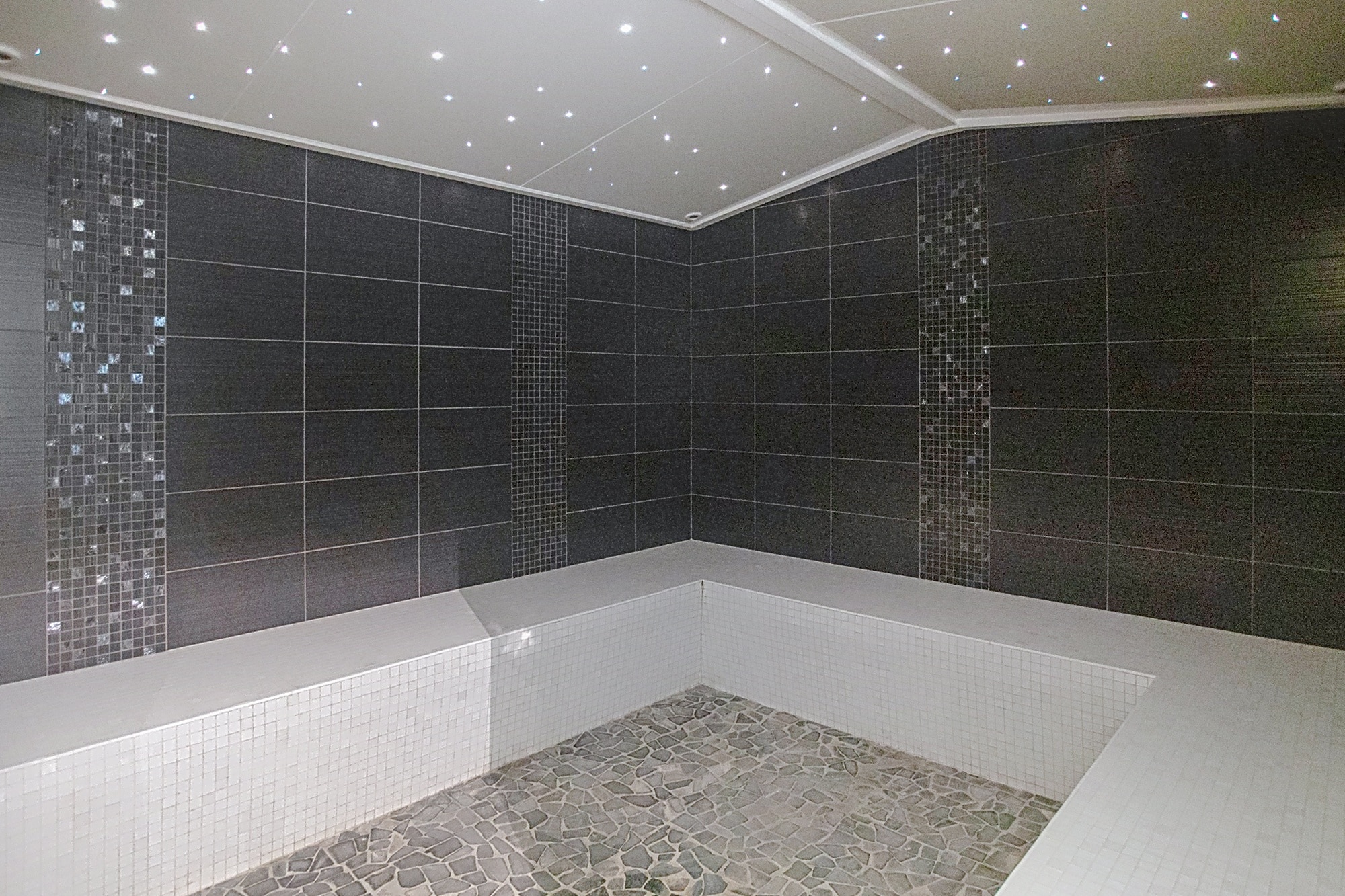 Hammam moderne avec murs en carrelage gris foncé, bancs blancs en mosaïque et plafond étoilé par des LED.