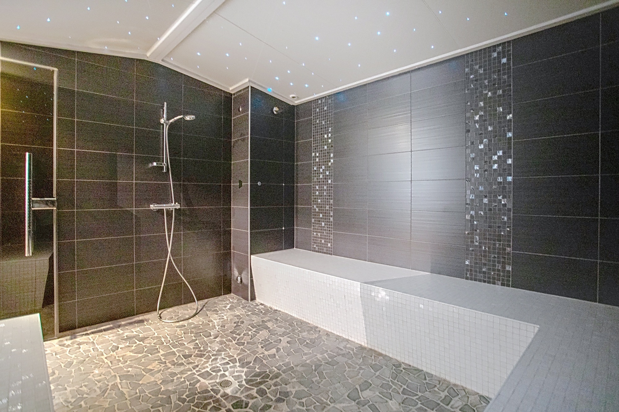 Hammam moderne avec carrelage gris, sol en galets, banc blanc et plafond ciel étoilé.