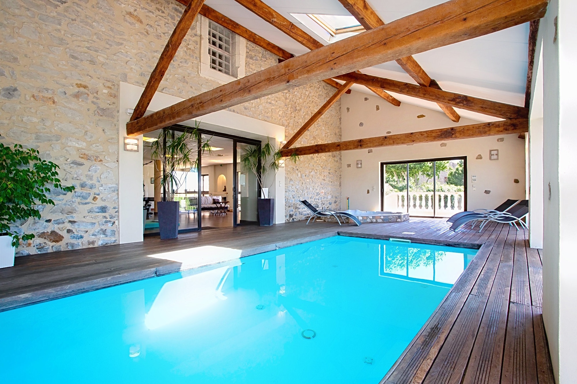 Piscine intérieure luxueuse avec murs en pierre apparente, charpente en bois et terrasse en bois exotique.