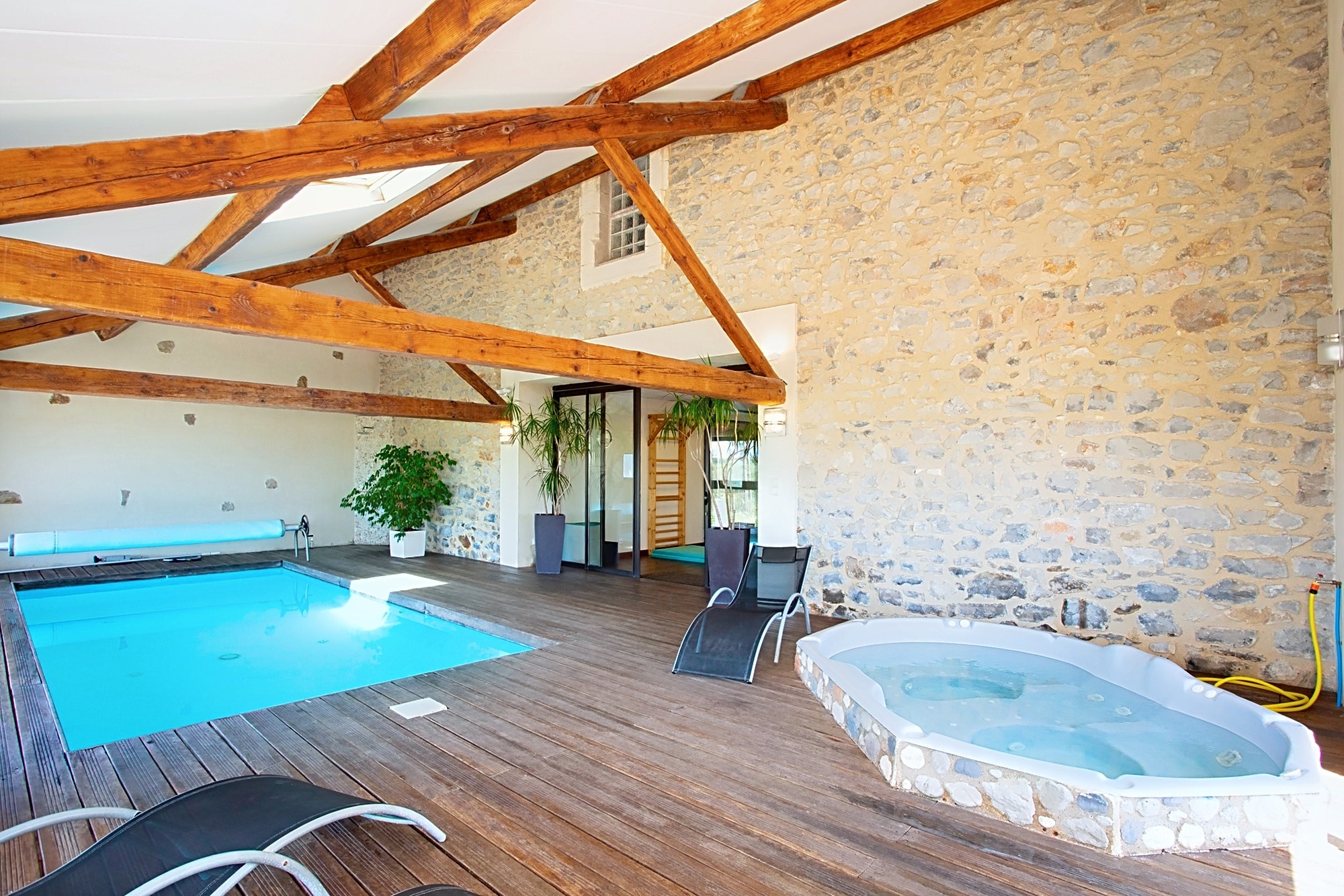 Piscine intérieure et jacuzzi dans un spa rustique avec poutres apparentes et mur en pierre naturelle.