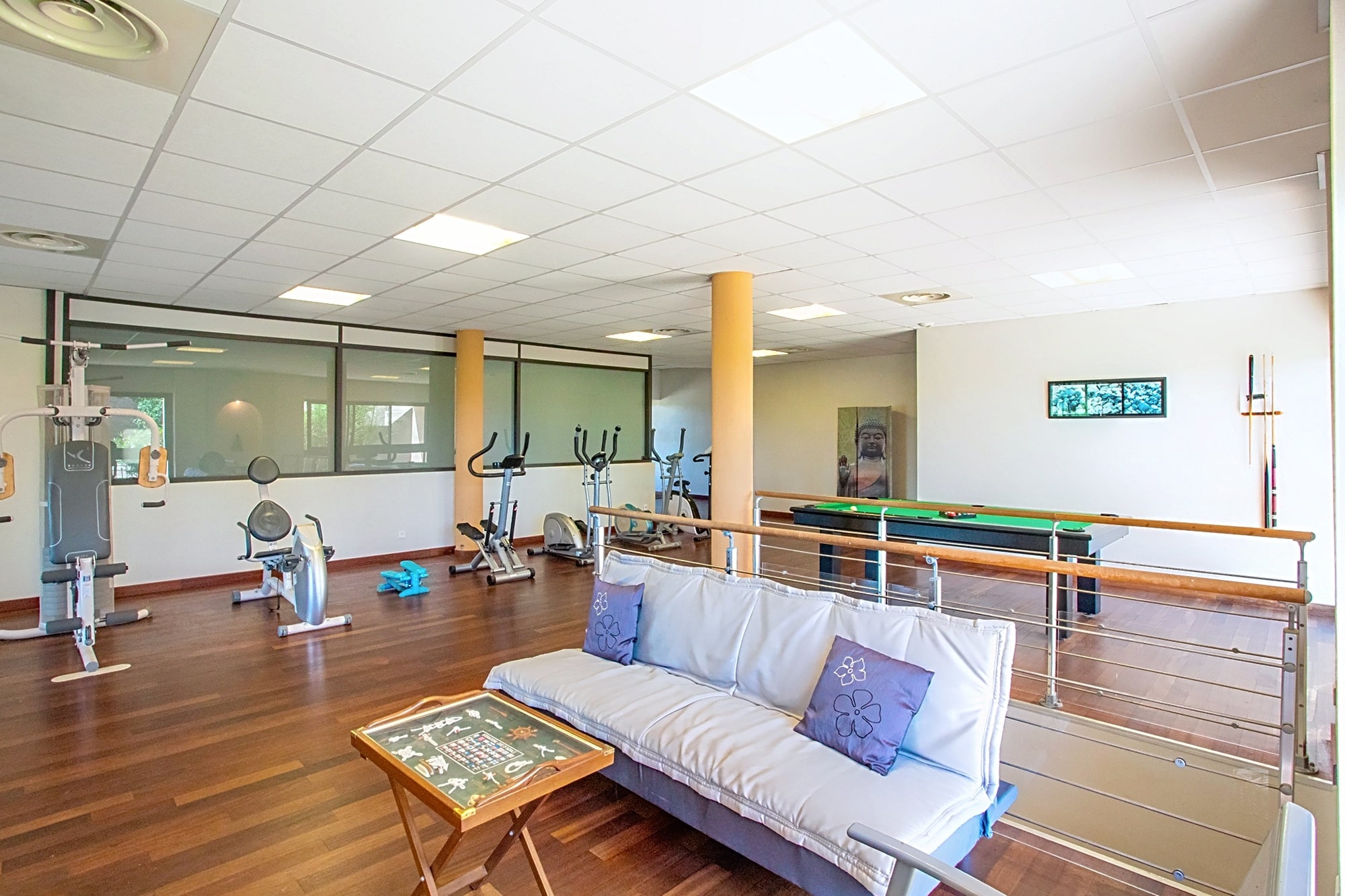 Salle de fitness lumineuse avec appareils de cardio, musculation, canapé et billard sur parquet en bois.